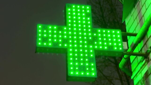 Green pharmacy cross glowing on street at night, LED diodes form bright matrix mounted on stone facade, vivid neon reflection.