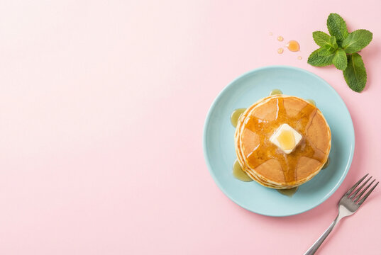 Top view of delicious fluffy pancakes stack with melting butter and sweet maple syrup on a pastel blue plate over a pink background with fresh mint leaves and a silver fork, breakfast food overhead.