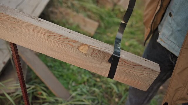 Hands fixing wooden beam outdoors, closeup of jacketed worker securing strap and checking fastener, carpenter adjusting timber plank on rural fence post, autumn grass background, methodical manual