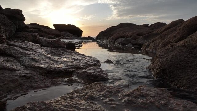 The sun rises through scattered clouds, over a rocky shore, casting a golden glow over the rocks onto the water's surface as the waters recede at Ramsgate, South Coast.
