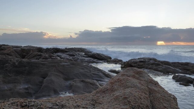 Sunbeams pierce through dramatic clouds over a rugged coastline as waves crash against the rocks, creating a serene yet powerful natural scene at Ramsgate, South Coast.