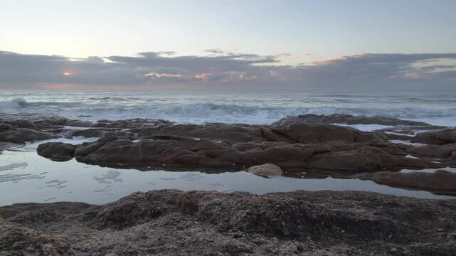 Gentle ocean waves roll onto a rugged coastline with tide pools reflecting the soft hues of the early morning sky at Ramsgate, South Coast.
