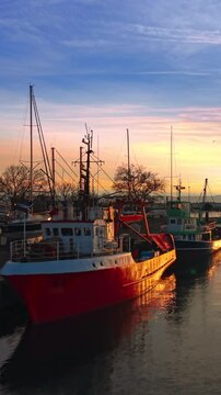 Red and white fishing vessel docked in a harbor with warm sunset light reflecting on the water. Fishing boat in port during golden hour. Vertical video.
