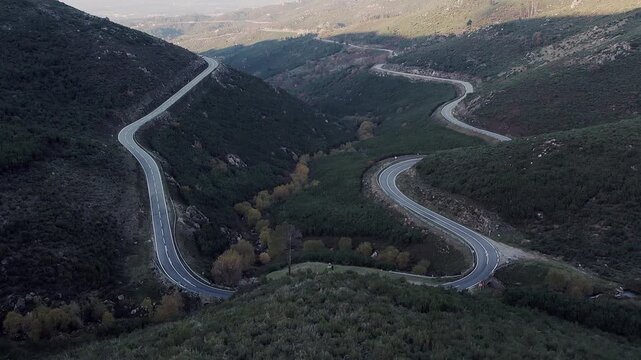 N338 mountain road with multiple switchbacks winding through steep valley in Serra da Estrela, Central Portugal, showing shrub vegetation, riparian trees and rugged high-altitude terrain, drone orbit