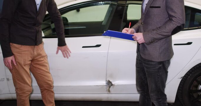 Driver explains accident details to insurance agent in garage closeup. Man inspector with clipboard interviews owner about vehicle crash at service station