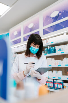 Professional female pharmacist wearing a face mask and white coat uses a digital tablet with a stylus to manage medical inventory and records at a pharmacy workstation.