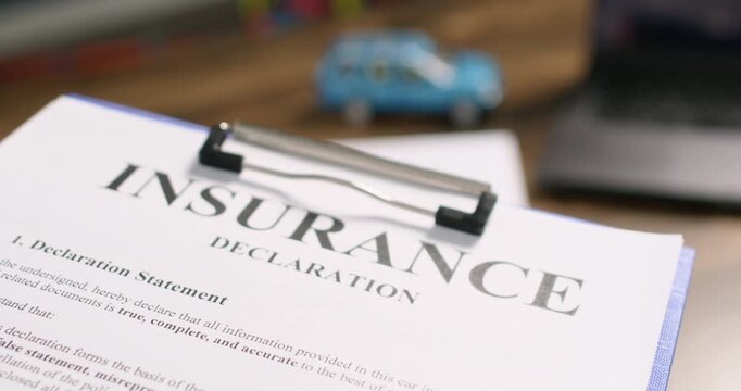Insurance declaration rests on clipboard near toy car and blurred laptop. Document shows printed policy text while supporting contract review for driver coverage