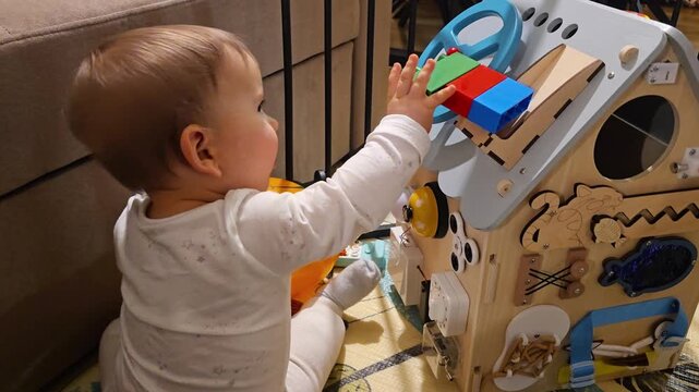 Little toddler playing with wooden busy board house educational toy developing fine motor skills and sensory exploration in a cozy home environment using Montessori method. 