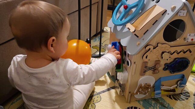 Little toddler playing with wooden busy board house educational toy developing fine motor skills and sensory exploration in a cozy home environment using Montessori method. 