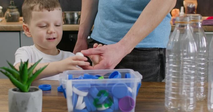 Father offers blue cap near box while smiling boy reaches from table. Child grabs plastic lid dropping piece into container carefully during recycling lesson