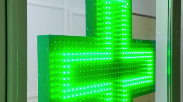 Green led pharmacy cross sign hanging, vibrant diode matrix pulses across suspended fixture, glass storefront reflection, clinical.