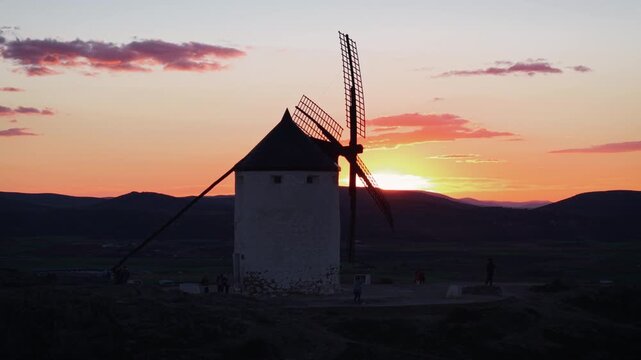 Iconic historic windmill in Consuegra, Spain, standing against a beautiful orange and pink sunset, with the sun disappearing behind the mountains of La Mancha
