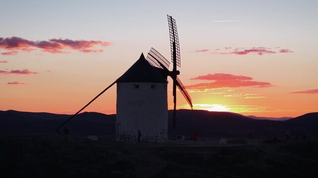 Traditional windmill from Consuegra, Spain, turning its blades during a beautiful orange sunset with mountains in the background and a plane flying in the sky
