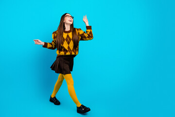 Naklejka premium Young woman in yellow argyle sweater and tights walking confidently on bright blue background, stylish casual fashion portrait showing joyful energy and retro-inspired outfit