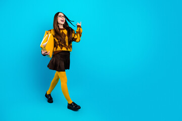 Fototapeta premium Happy young student walking with yellow folder and tights on blue background wearing argyle sweater and glasses, stylish schoolgirl portrait full length with confident joyful expression