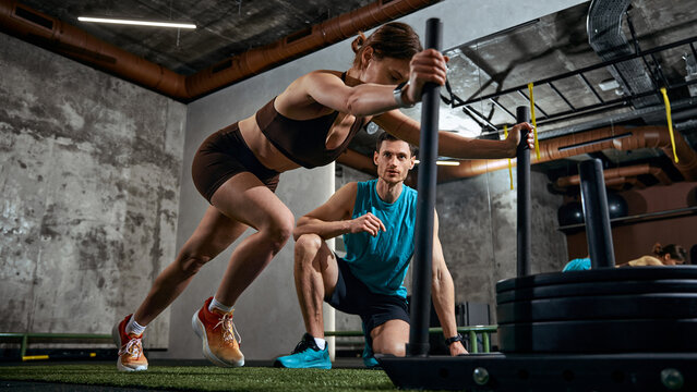 Woman pushing weighted sled while man supervising training session in gym. Concept of strength training promo, fitness ads campaign, athletic branding and performance coaching service marketing