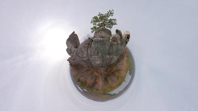 Man on top of a cliff. View of a small planet.  A granite cliff with a pine tree growing on its summit and a panoramic view of a mixed forest and lakes in the distance.  360 degree camera. 