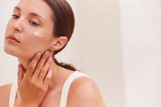 Skin care. Woman applying face cream with a visible white swatch on her cheekbone, touching her jaw gently against a soft neutral background.