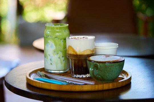 Tray with clay mug of cappuccino, glass latte, and matcha latte on small wooden table in Vietnamese coffee shop, beautiful bokeh background.