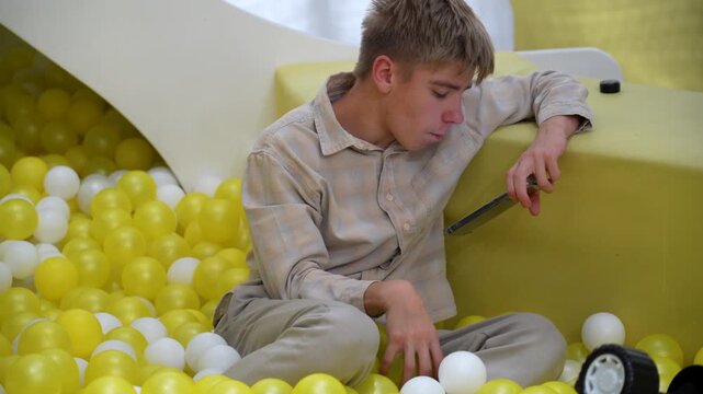 Teenager playing with ball pit and smartphone, lost in thought amidst sea of colorful spheres, reflecting on digital connection.
