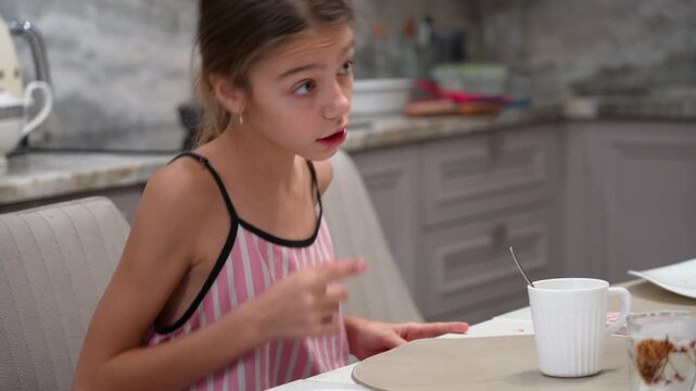 Young girl with braided hair expresses frustration while sitting at kitchen table with cup and saucer, then appears contemplative.