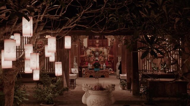 Traditional Vietnamese temple courtyard at night in Hanoi with illuminated paper lanterns hanging from tree and peaceful altar inside building for worship and prayer