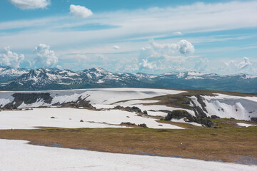 Scenic alpine landscape with sunlit snowy field on stony hill with view to big mountain range with forest and snow far away under cloudy sky. Stone outcrops among snows in sunlight in high mountains. © Daniil