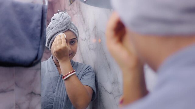 Young female gently dabbing makeup puff on face during her morning makeup routine, wearing bathrobe in the modern bathroom.