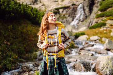 A woman enjoys her time exploring the mountains with a backpack. She stands near a waterfall and looks happy while surrounded by green plants and rocks. Hiking concept. © maxbelchenko