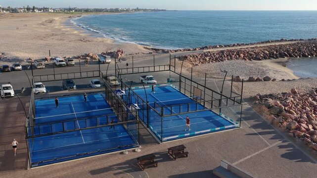 People playing padel tennis on outdoor blue courts by ocean in Namibia, with sandy beach and coastal town visible along shoreline of Africa from aerial angle