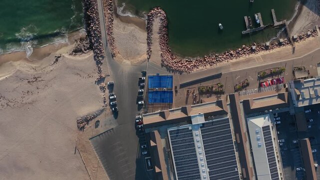 People playing padel on blue court at a waterfront resort with solar panels on the roof, next to sandy beach and marina with boats on Atlantic Ocean in Namibia