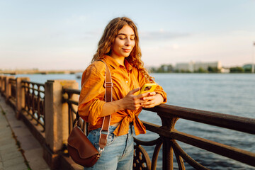 A woman stands by a bridge, looking at her phone. The sun sets behind her, bathing the landscape in warm light. She is relaxed and dressed in casual clothes. © maxbelchenko