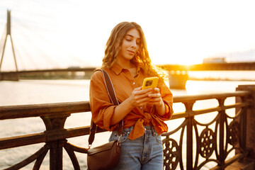 A woman stands by a bridge, looking at her phone. The sun sets behind her, bathing the landscape in warm light. She is relaxed and dressed in casual clothes. © maxbelchenko