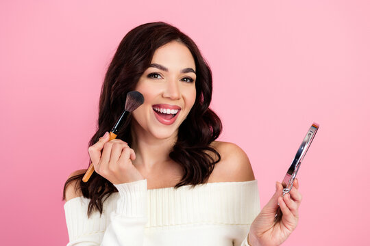 Young woman applying makeup with brush and mirror, smiling beauty portrait on pink background conveying joyful self-care