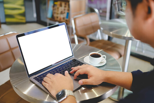 A man is typing on a laptop with a blank white mockup screen while sitting at a metallic cafe table with a cup of black coffee.