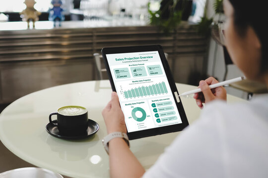 A close-up view of a person using a stylus to analyze a sales projection overview dashboard on a tablet while sitting at a cafe table with a matcha latte.
