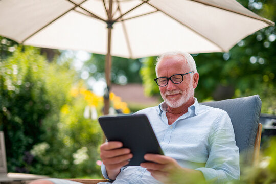 A happy elderly man sitting in a garden under a sun umbrella, reading from a tablet. Edited AI generated image
