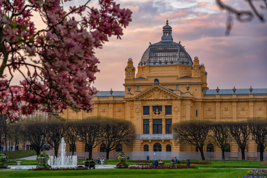 Historic Art pavilion building in Zagreb surrounded by blooming magnolia trees and spring park landscape at sunset, with soft pastel sky