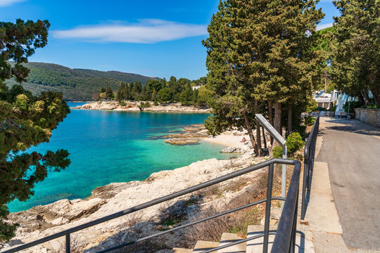 Scenic Adriatic coastline in Rabac town with turquoise crystal clear water, rocky shore and pine trees, with a coastal walkway and railing overlooking the bay on a sunny day