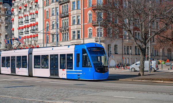 A modern blue and white tram travels along a historic street lined with elegant architecture and red-awning buildings in Stockholm, Sweden.