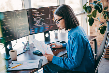 Female programmer in a modern loft office analyzes code and documents at dual monitors