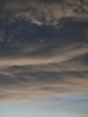 Fototapeta premium Soft moody sky filled with layered dark gray and warm beige wispy clouds stretching across the frame, with a narrow pale blue band visible near the horizon.