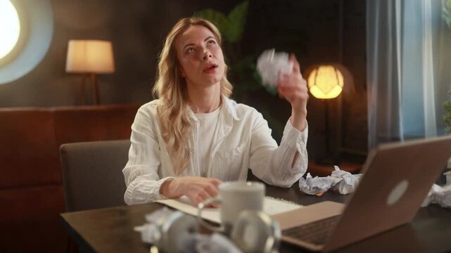 Young woman surrounded by crumpled papers, sitting at home and struggling to come up with ideas, appearing mentally exhausted and overwhelmed, facing creative block or burnout in a quiet workspace