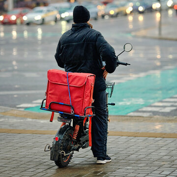 Food delivery. Rear view of food delivery courier standing with an electric moped and large red thermal bag on wet city sidewalk during rainy evening with blurred traffic lights in the background