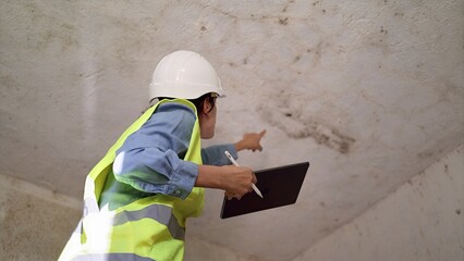 Building inspector wearing hard hat and safety vest evaluating ceiling damage, pointing at dark mold growth and water stains during a property inspection with a digital tablet © NJF