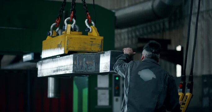 Rear view of a factory worker using a magnetic lifter to move a heavy steel block in a workshop