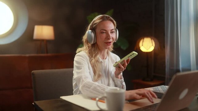 Young woman working on her laptop while recording voice messages, speaking into her smartphone as she organizes tasks and ideas, using an audio planner during a productive remote work session at home
