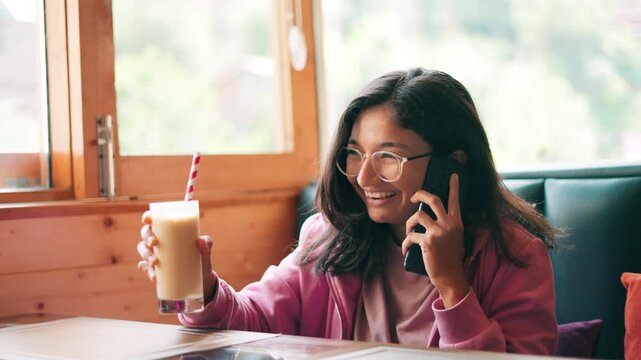 Cheerful Indian woman talking on call and drinking cold coffee in cafe at Manali, India. Laughing young woman on cell phone in a cafe. Female enjoys morning coffee.
