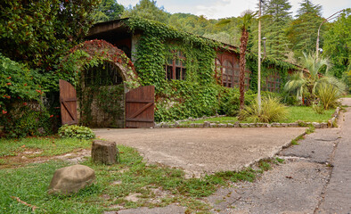 Naklejka premium An old, ancient stone house, completely immersed in greenery, with wooden gates, in countryside on warm evening. Not AI.