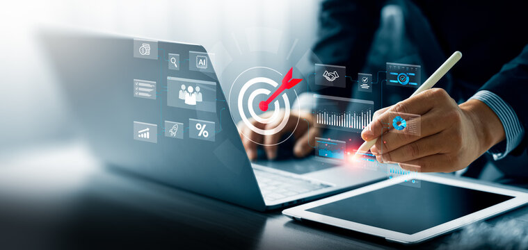 Business professional using stylus on tablet with laptop and digital target icon interface, showing analytics dashboards, charts, and business data for strategy planning and performance tracking